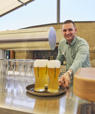 Mitarbeiter des Blockbräu serviert mehrere frisch gezapfte, goldgelbe Biere mit Schaumkrone an der Bar auf der Hafenterrasse.