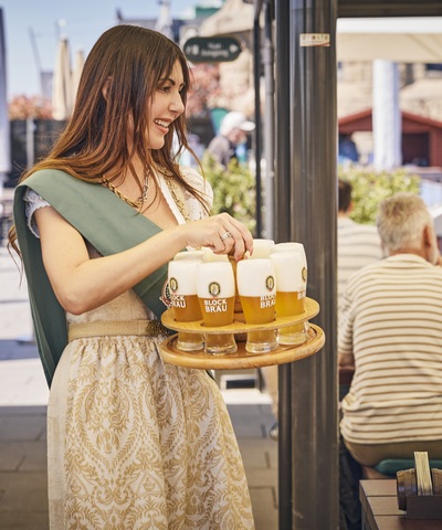 Mitarbeiterin im Dirndl serviert mehrere frisch gezapfte Blockbräu Biere auf einem großen Holztablett an Gäste auf der sonnigen Hafenterrasse.