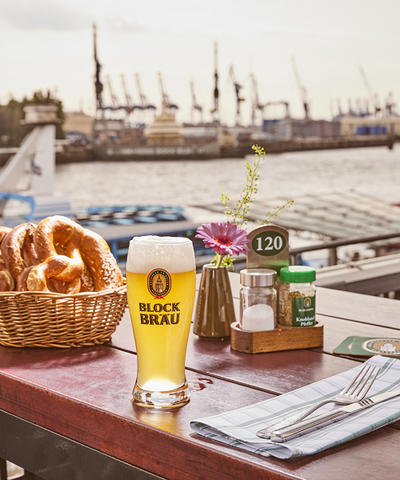 Frisch gezapftes Blockbräu Bier und ein Korb mit Brezeln auf einem gedeckten Tisch der Hafenterrasse. Im Hintergrund sind die Elbe und die Hafenkräne des Hamburger Hafens zu sehen.