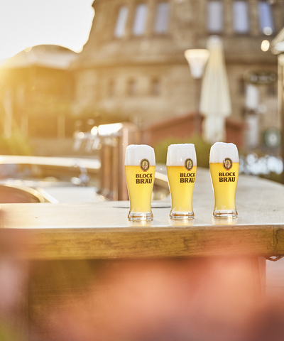 Drei frisch gezapfte, goldgelbe Blockbräu Biere mit kräftiger Schaumkrone stehen im warmen Abendlicht auf der Theke der Hafenterrasse.