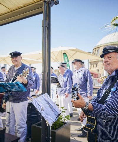 Chor in maritimer Kleidung musiziert bei Sonnenschein auf der Blockbräu Hafenterrasse mit Gitarren und Akkordeons. Im Hintergrund ist der markante Turm der Hamburger Landungsbrücken zu sehen.