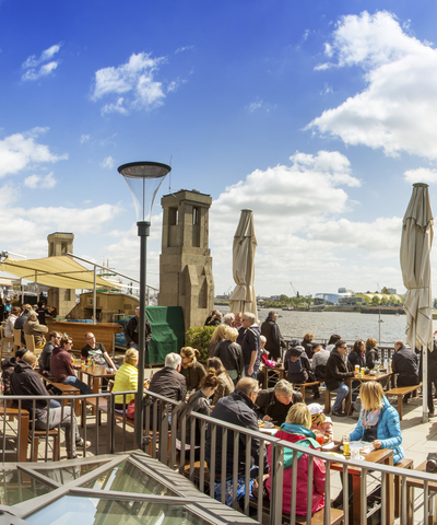 Blockbräu Hafenterrasse bei sonnigem Wetter mit zahlreichen Gästen an Holztischen. Die Terrasse bietet einen weiten Blick über die Elbe und den Hamburger Hafen.