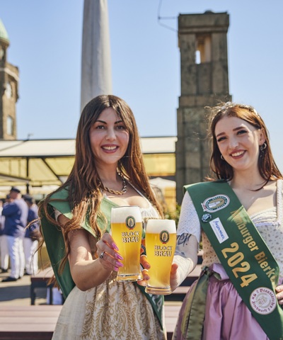 amburger Bierprinzessin und Hamburger Bierkönigin 2024 präsentieren zwei frisch gezapfte Blockbräu Biere auf der sonnigen Hafenterrasse.