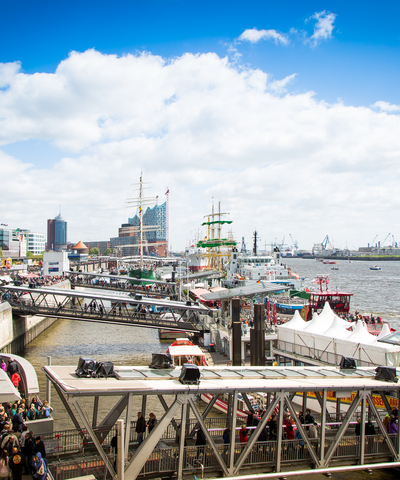 Panoramablick von der Blockbräu Hafenterrasse über die Hamburger Landungsbrücken und die Elbe. Im Hafen liegen mehrere Schiffe, im Hintergrund sind die Elbphilharmonie und zahlreiche Hafenkräne zu sehen.